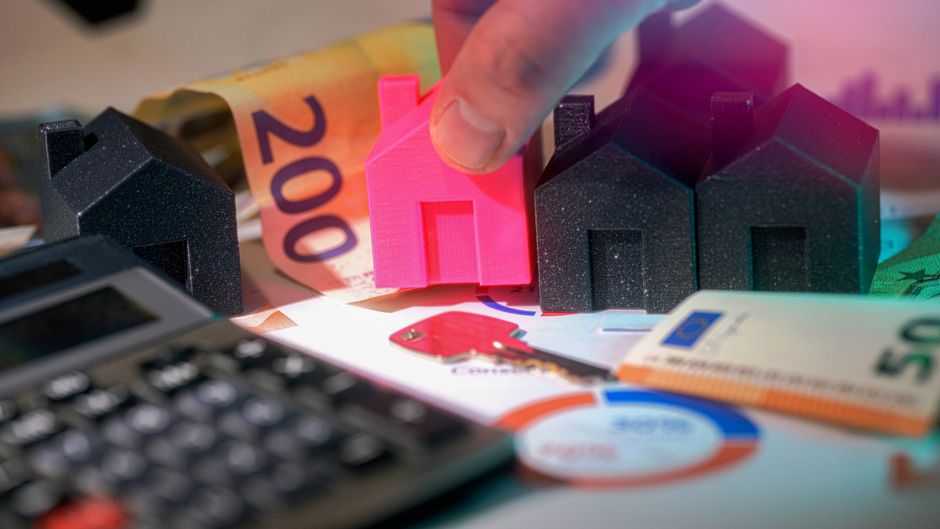 A close-up view of a person's hand placing a small, bright pink model house among three black and one pink scaled-down houses. The scene is set on a table cluttered with various items including a calc