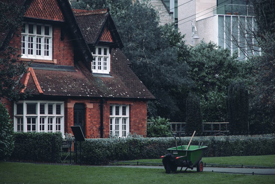 A traditional red brick terraced home with a sloped tiled roof and white-framed windows, situated in a landscaped garden. The garden features neatly trimmed hedges bordering a lawn area with lush, gre